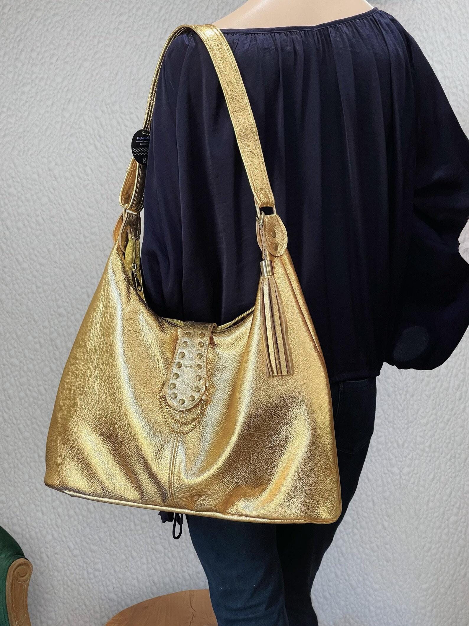 Gold metallic leather handbag worn by a person against a neutral background. leather tassel and brass hardware