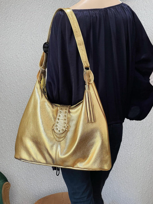 Gold metallic leather handbag worn by a person against a neutral background. leather tassel and brass hardware