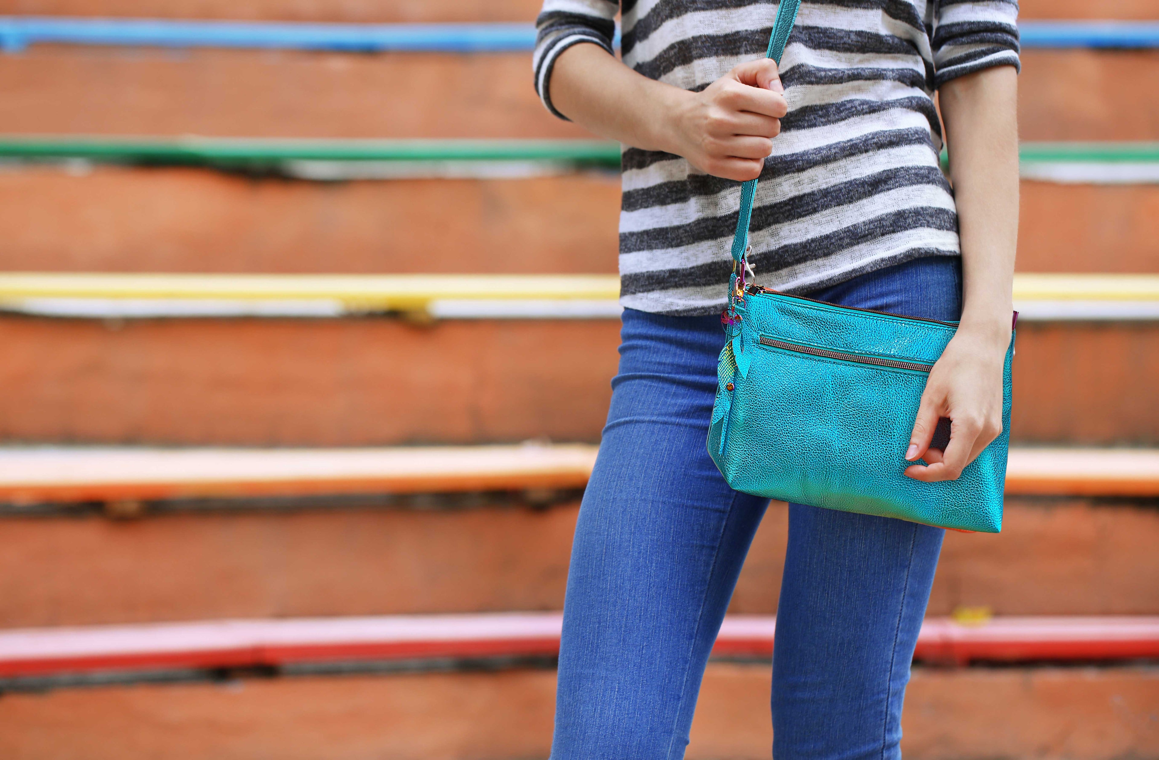 Person holding a turquoise clutch bag with a striped shirt and blue jeans against a wooden bench background