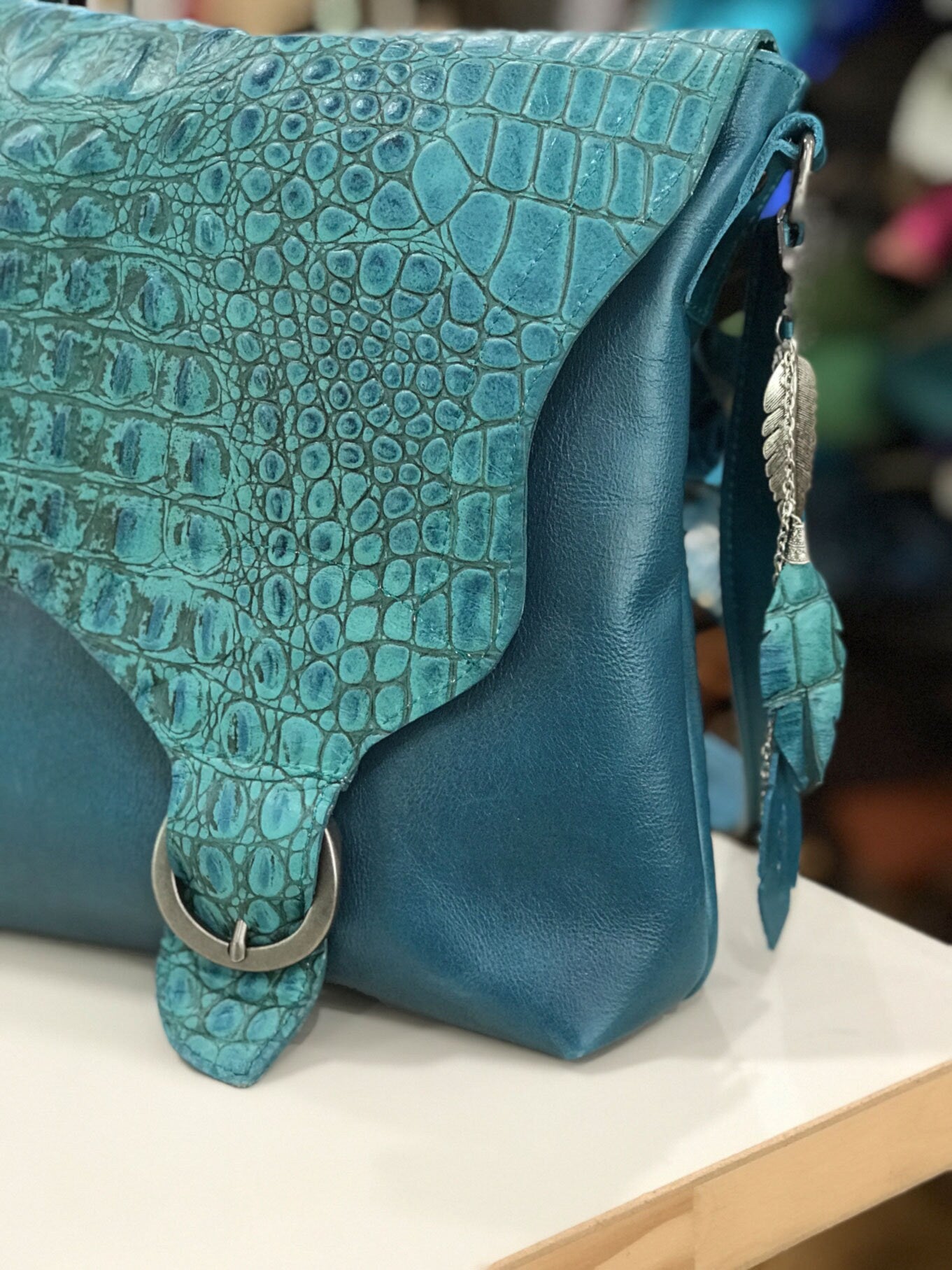 Close up of Teal handbag with crocodile texture flap and buckle and leather metal bag charm adornment