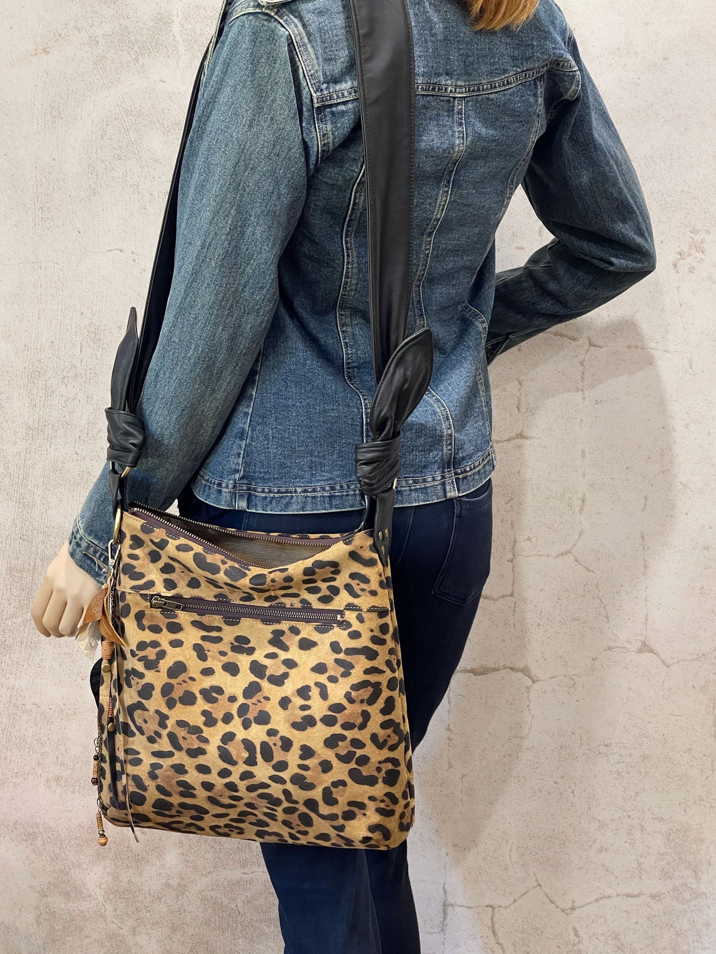Person wearing a leopard print bag over their shoulder with outside zipper pocket and black strap and top zipper closure.