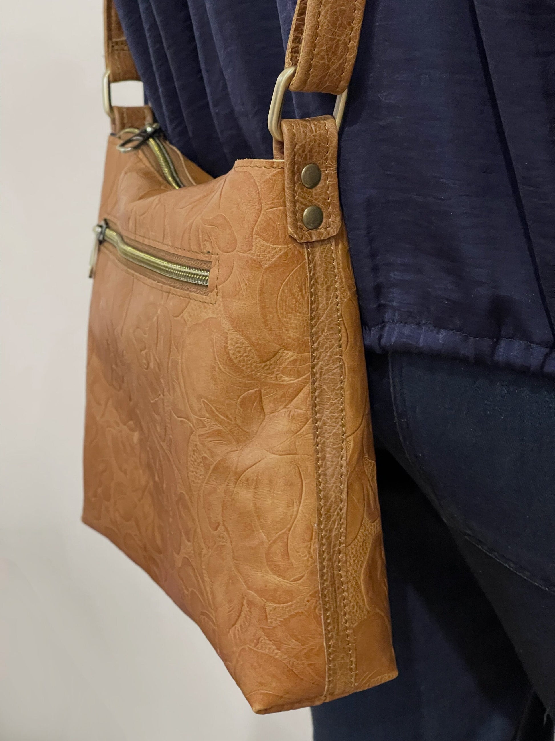 close up of side of tan leather handbag with floral patterns worn by a person in a navy jacket.