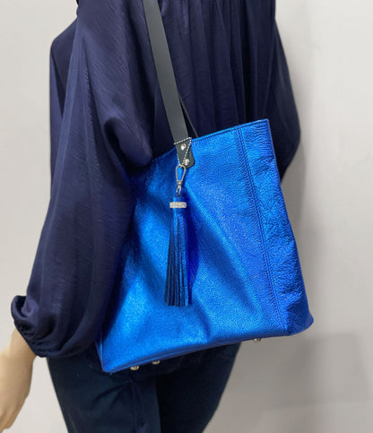 cobalt Blue metallic leather tote bag with long wearing black leather handles and a blue matching tassel held by a person wearing a dark blue outfit.