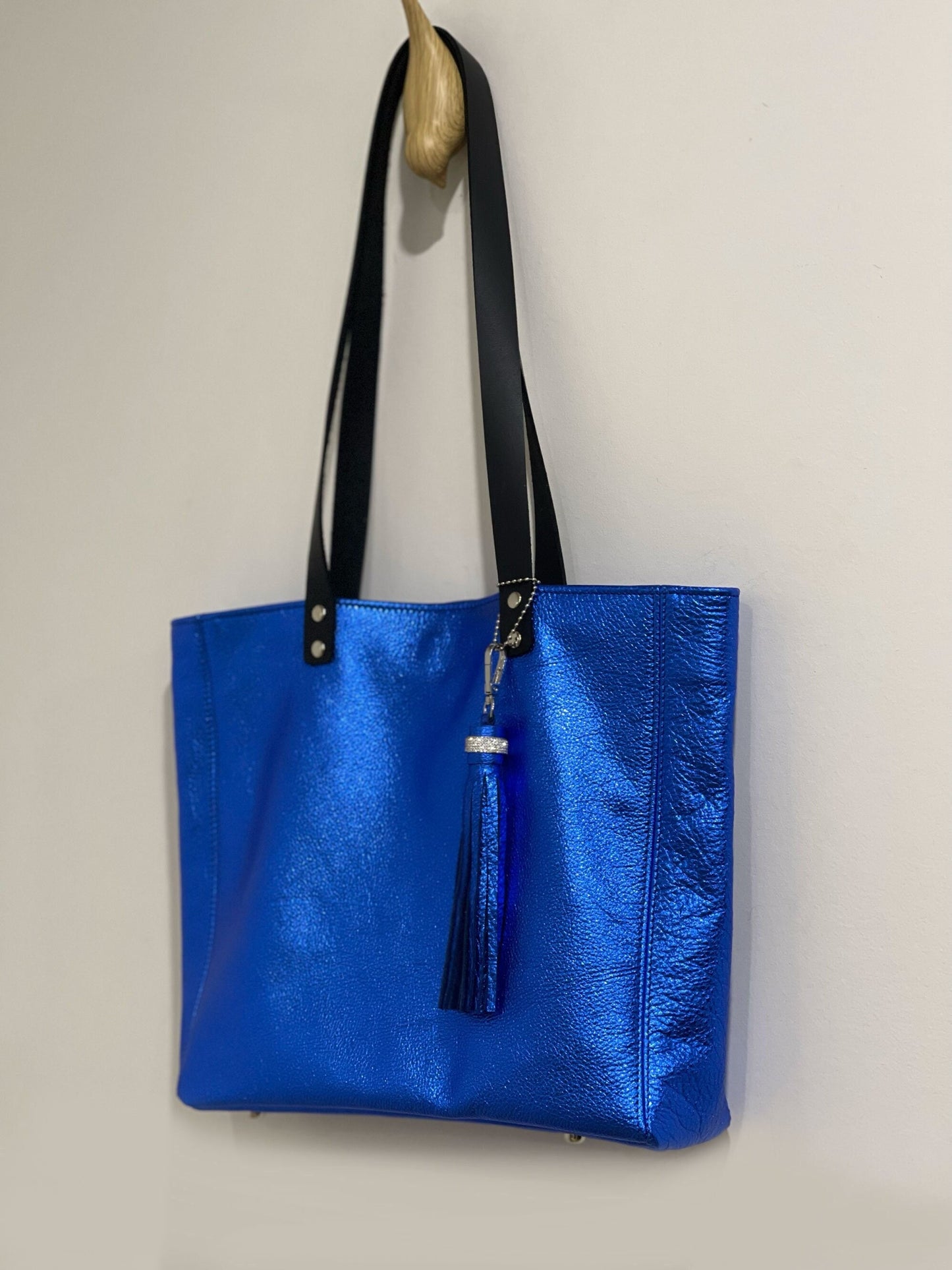 Cobalt Blue tote bag hanging on a hook with strong long shoulder straps in black leather and with a cobalt matching tassel on a beige wall