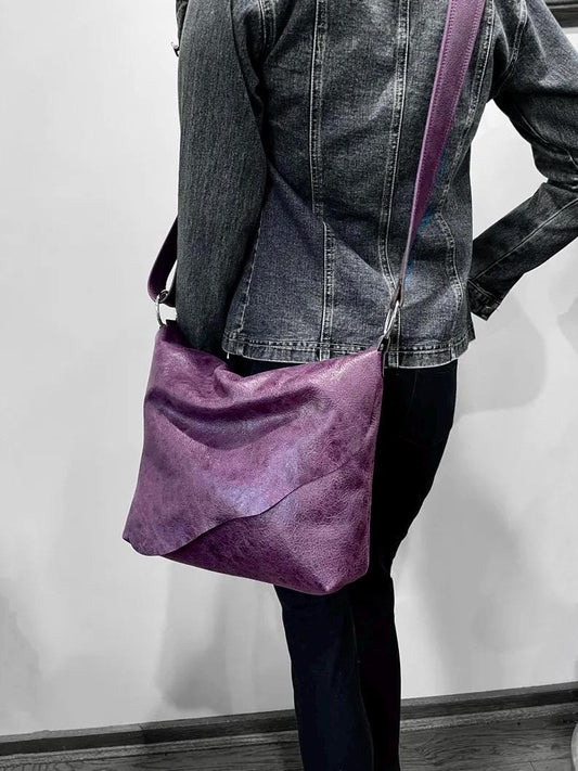 Person wearing a denim jacket with a distressed vintage purple shoulder bag with irregular long flap on a plain background