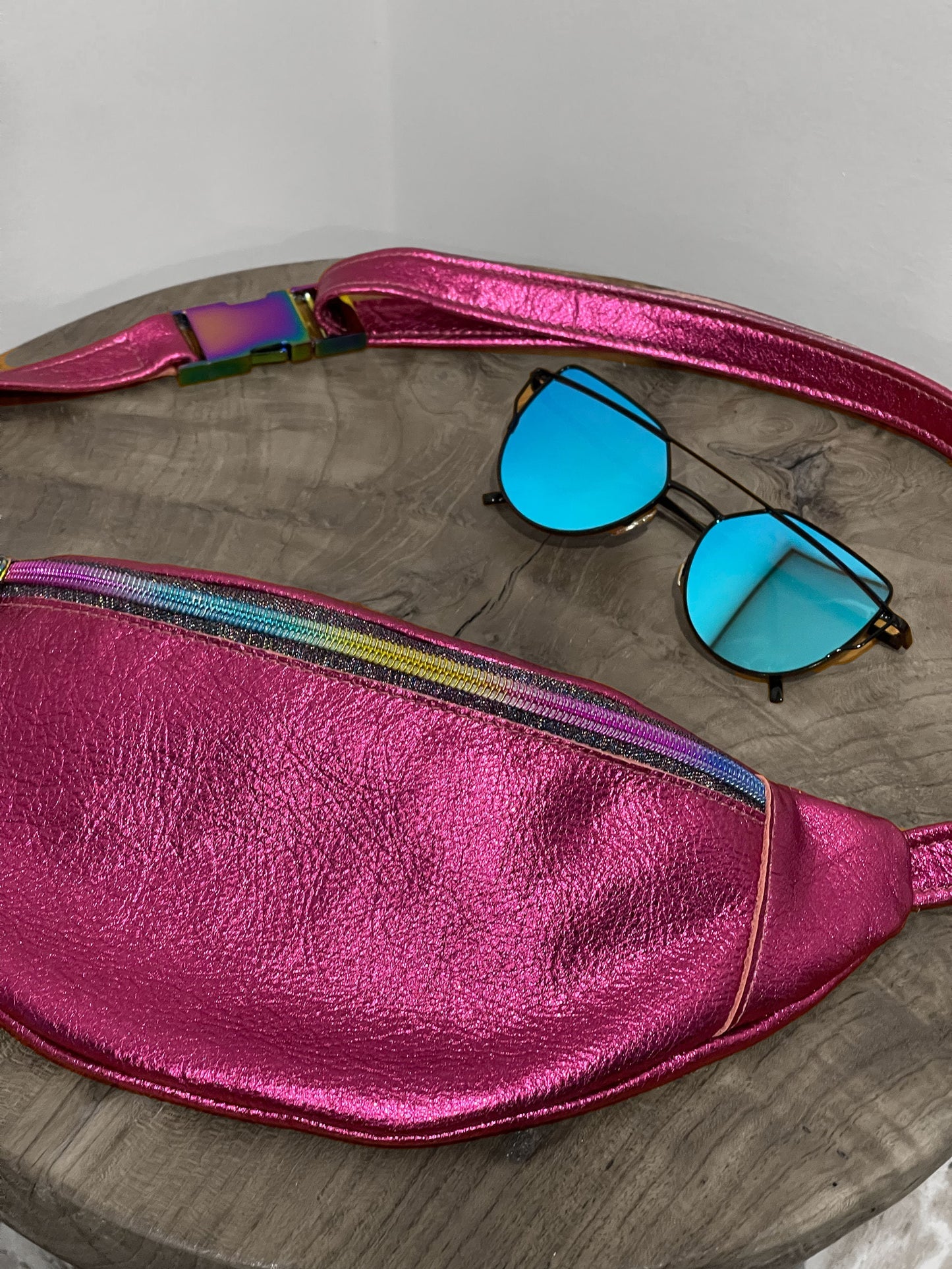 Pink metallic fanny pack with colorful strap and blue sunglasses on a wooden surface.