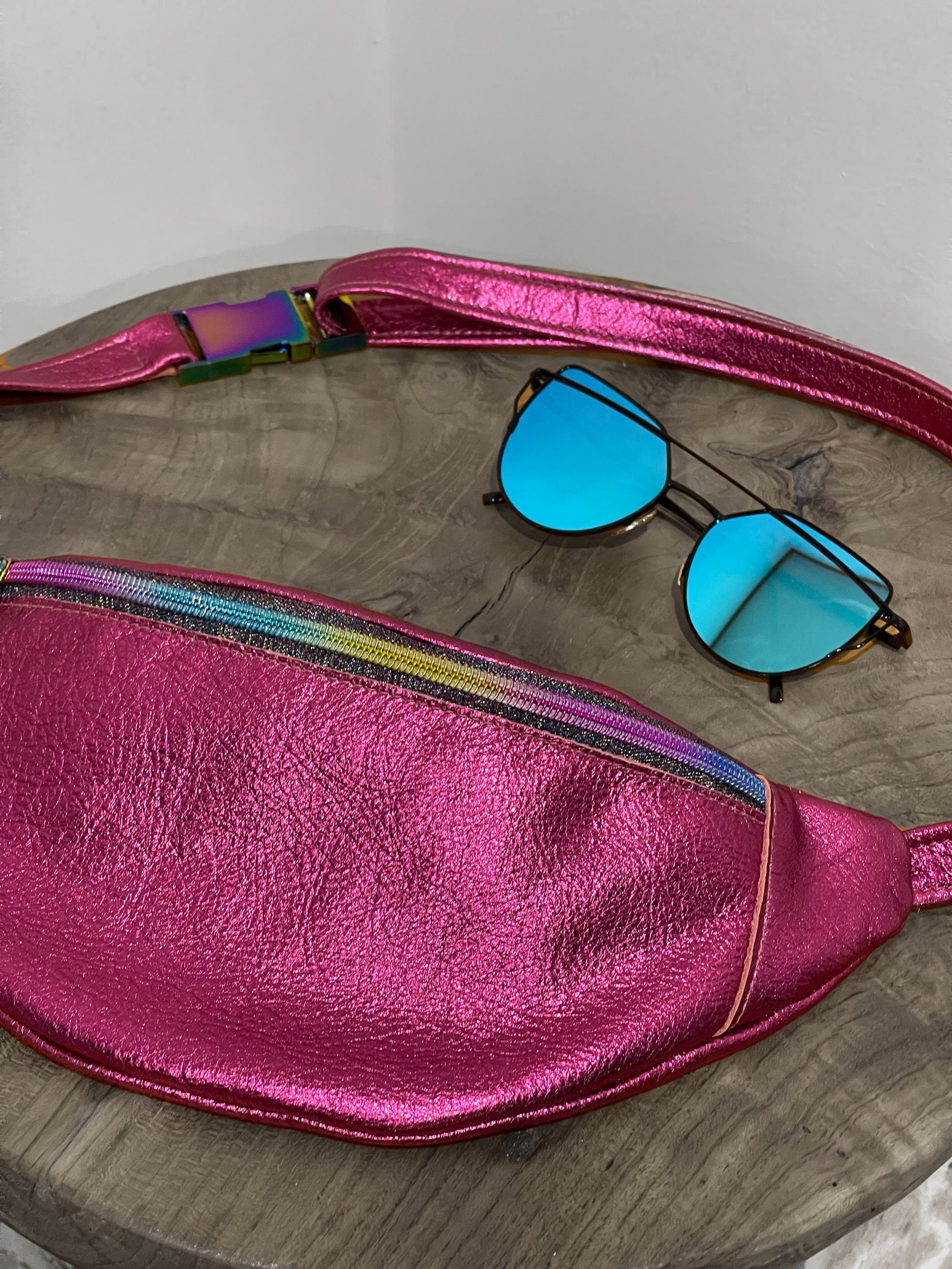 Pink metallic fanny pack with colorful strap and blue sunglasses on a wooden surface.