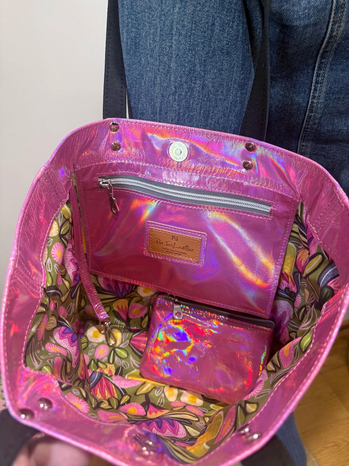 Holographic pink bag with floral interior, magnetic closure and strong riveted black leather straps worn over shoulder.