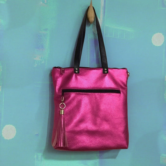 Pink tote bag with black accents and large pink tassel on the zipper hanging on a hook against a light blue wall.
