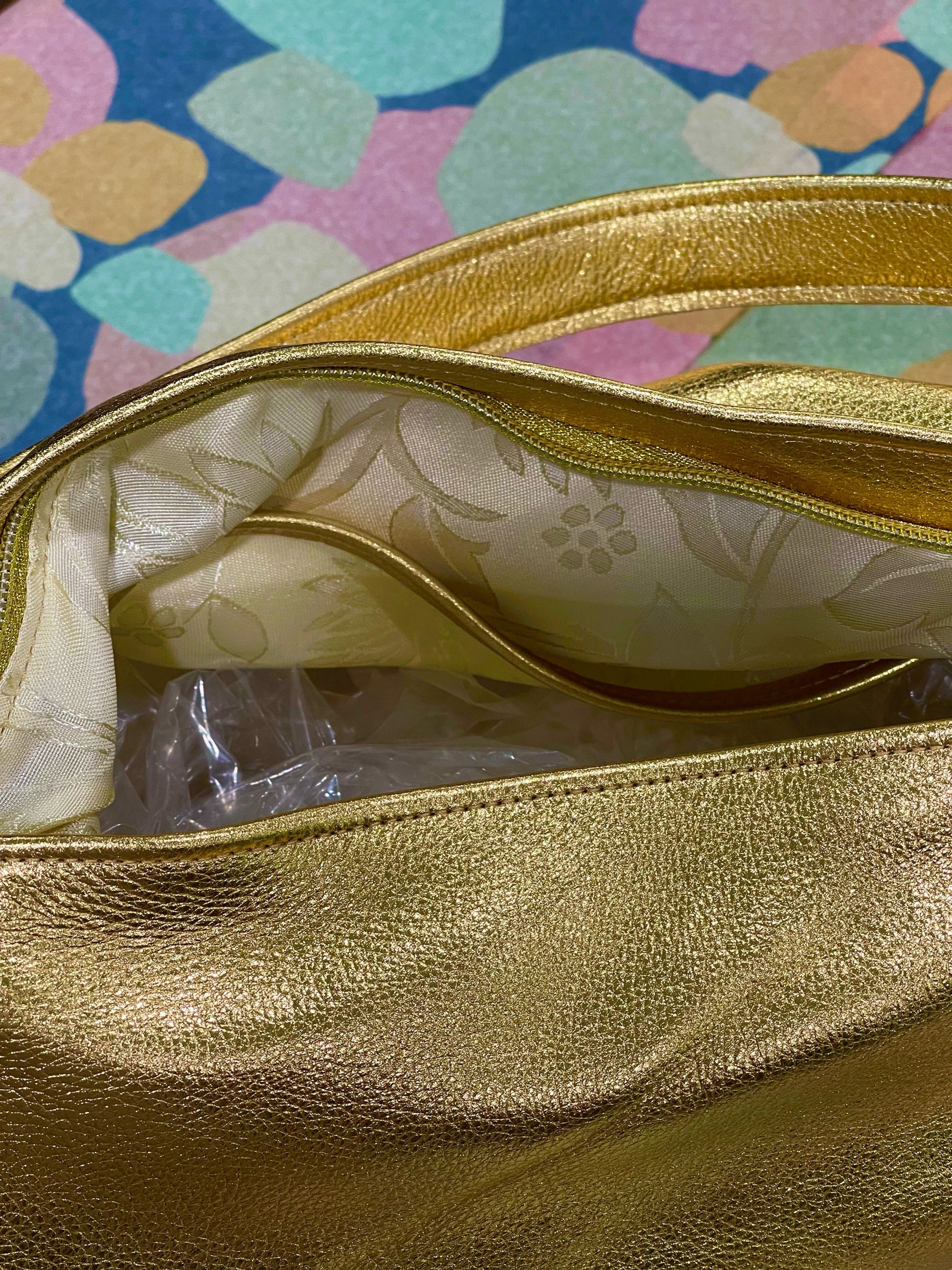 close up of a Gold handbag with floral lining and zipper closure set down into bag on a colorful patterned background