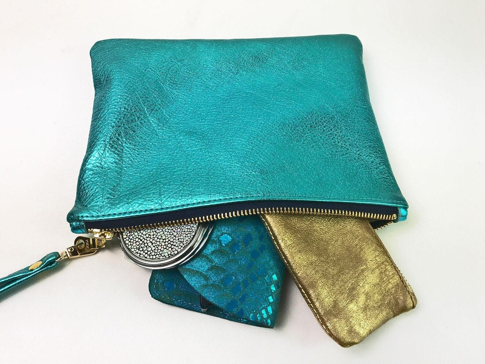 metallic teal clutch bag showing contents that fit the purse including makeup mirror and zipper pouch and sunglasses case.