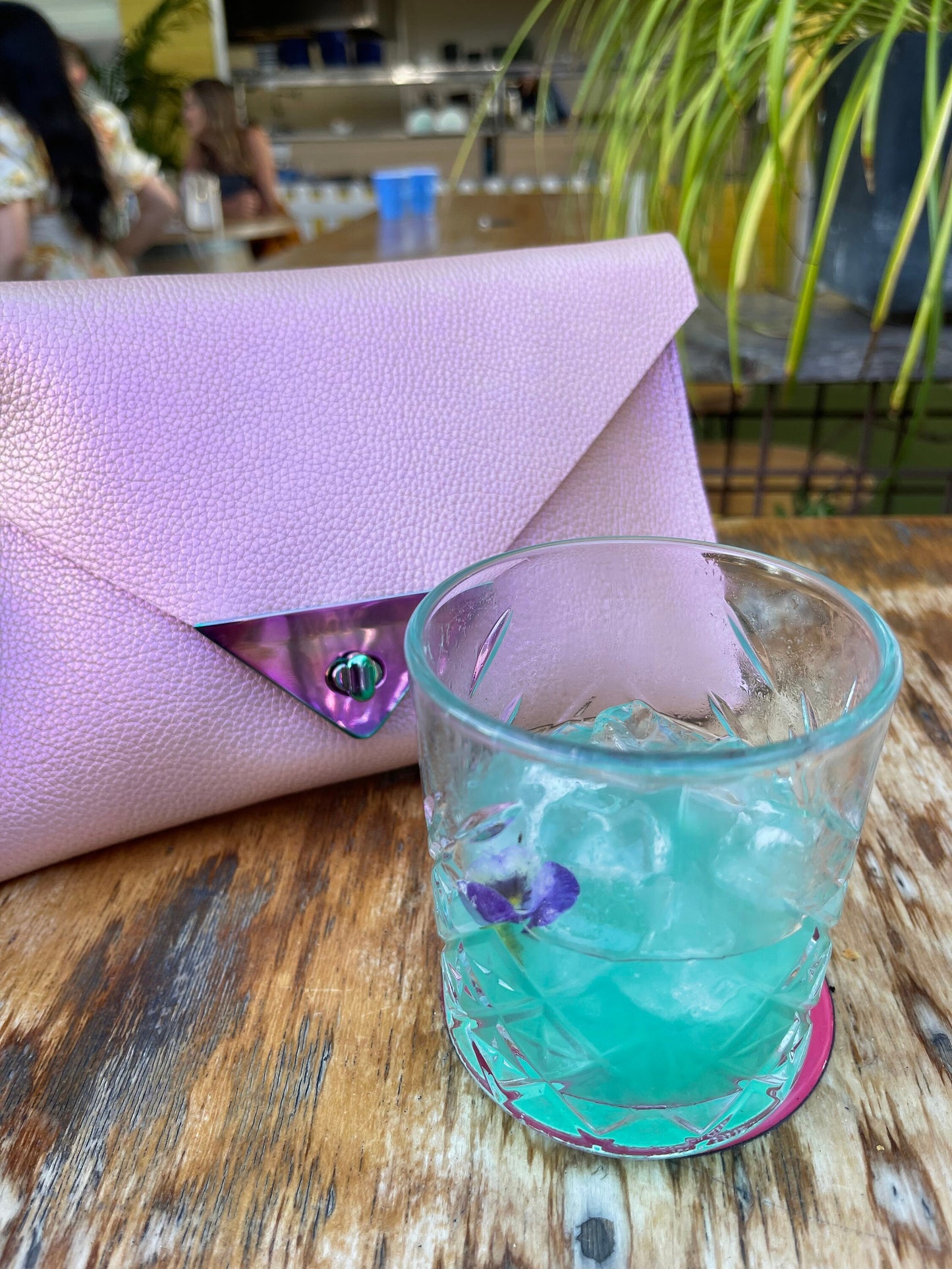 Blue cocktail in a glass with ice and a purple flower, next to a pink clutch with metallic pink clasp closure on a wooden table.