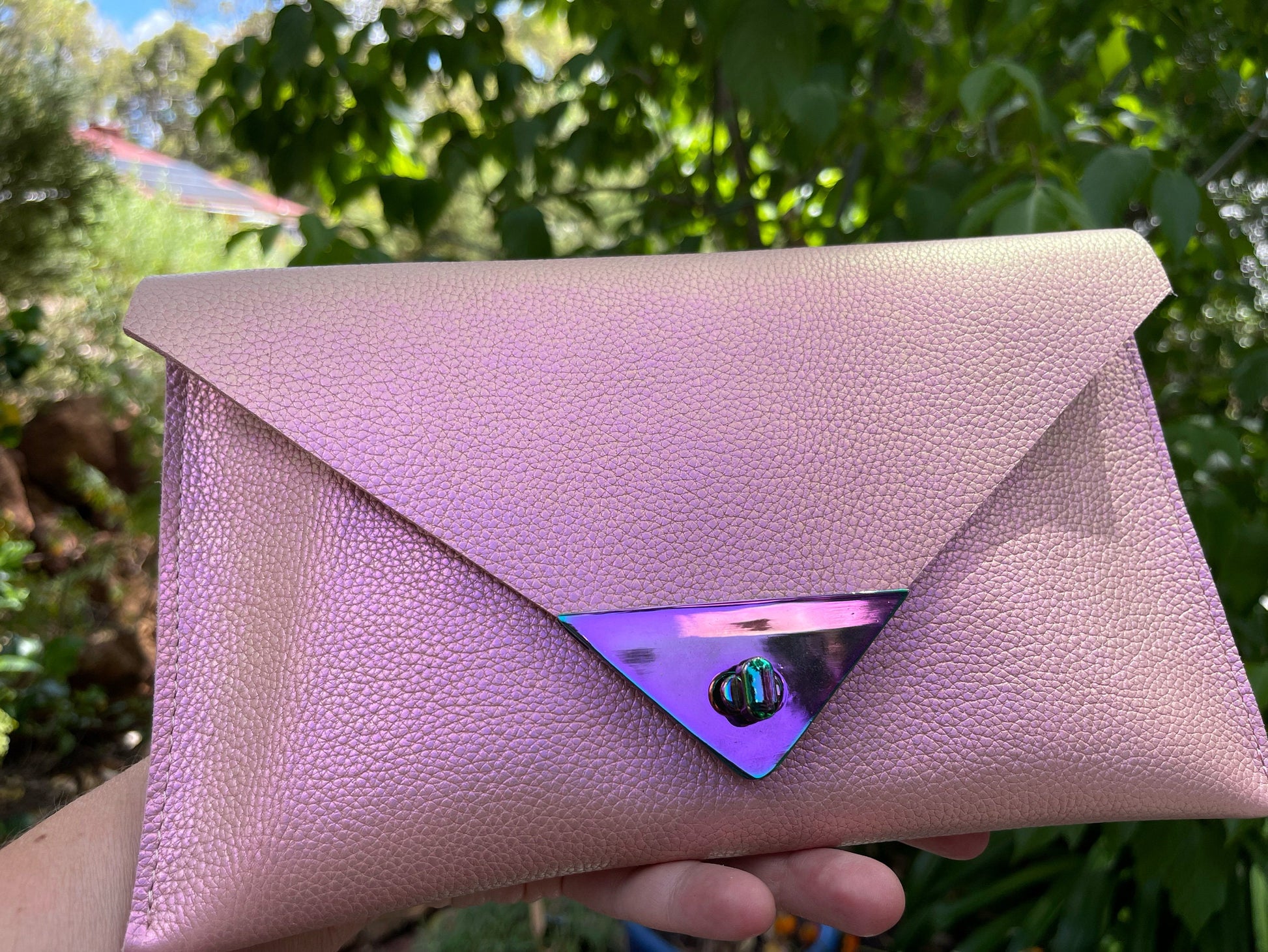 Pink clutch bag with a reflective purple triangle held in front of a green, leafy background