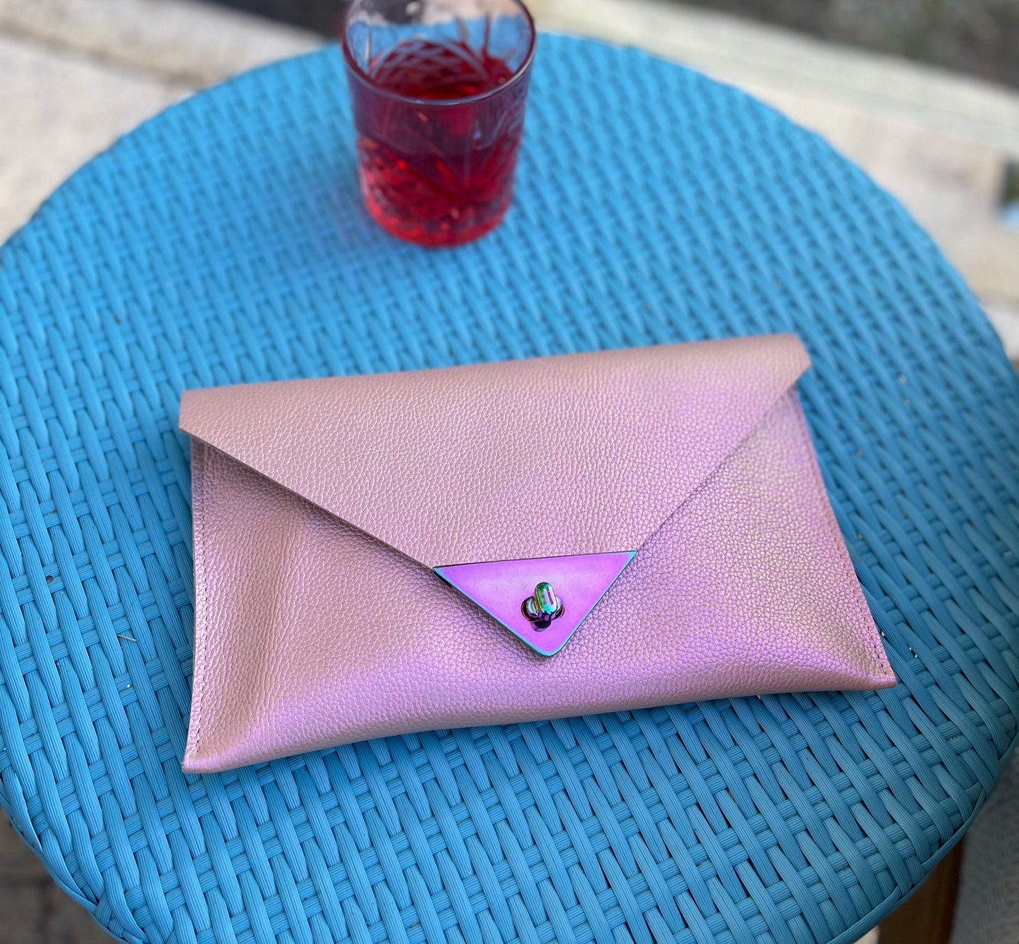 Pink envelope clutch on a blue wicker table with a glass of red liquid.