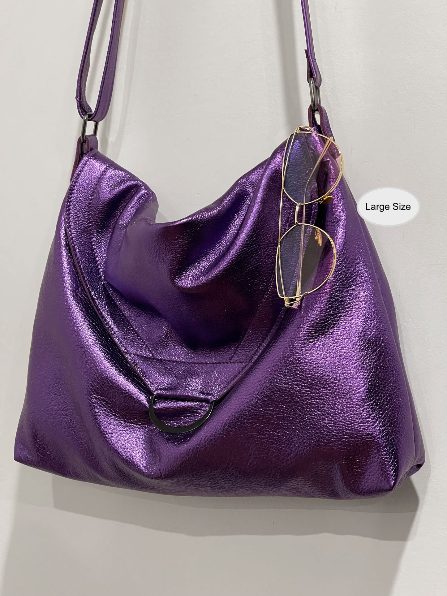 Purple handbag with flap close and large D ring and sunglasses hanging on edge on a light gray background