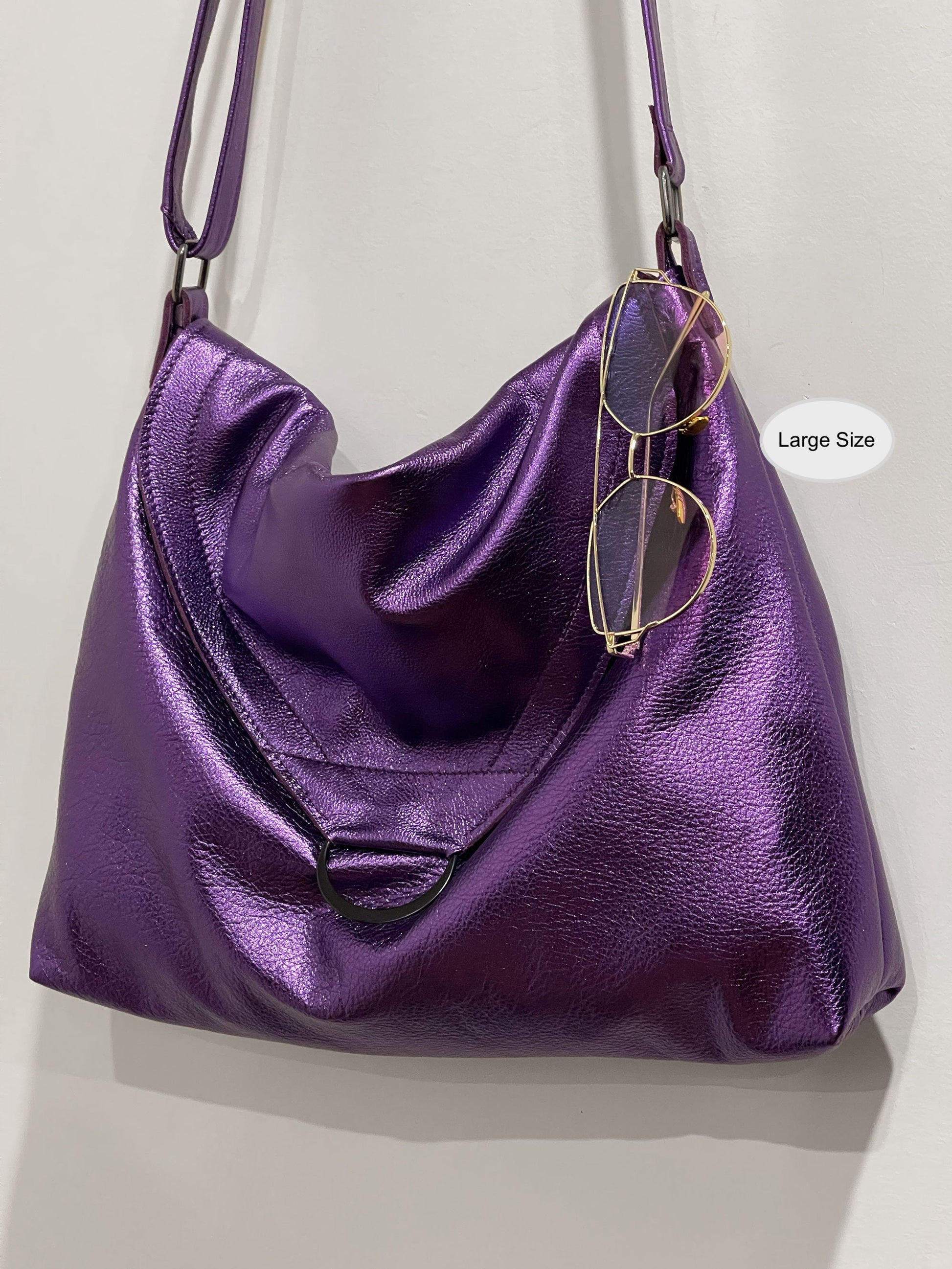 Purple handbag with flap close and large D ring and sunglasses hanging on edge on a light gray background