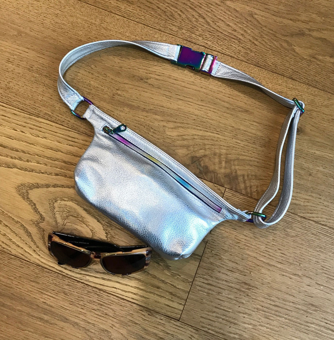 Silver metallic waist bag with a strong clasp on the strap and rainbow coloured zipper on a wooden surface