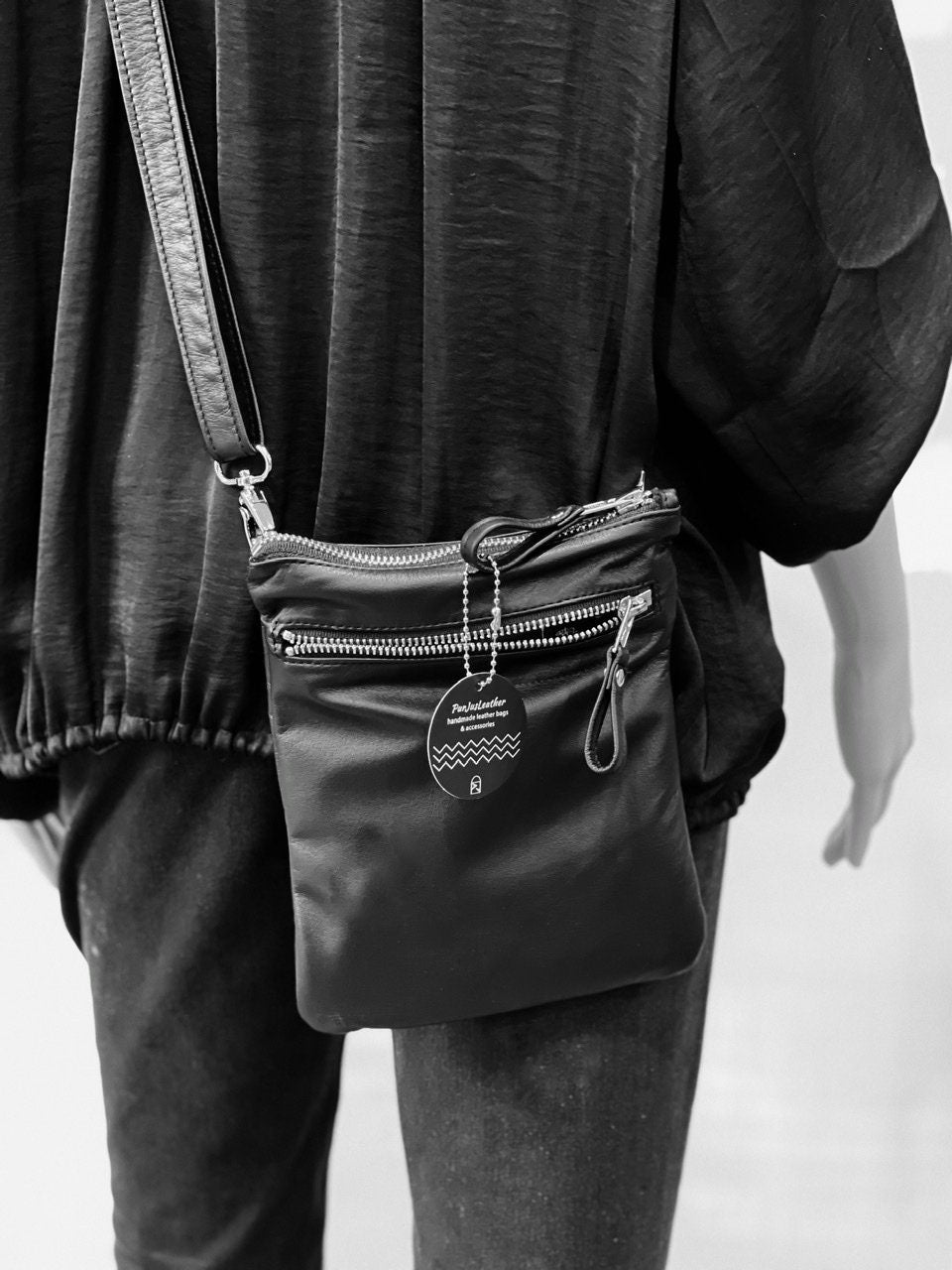 Person wearing a black crossbody bag with wide outside zipper pocket and leather pull with a visible brand tag on a light background