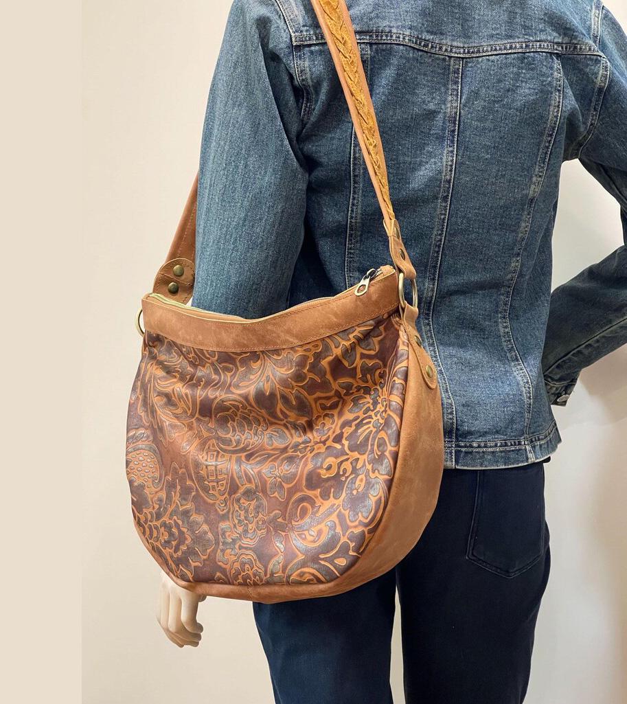 Model wearing a brown leather bag on shoulder with floral tooled patterns and tan leather sides