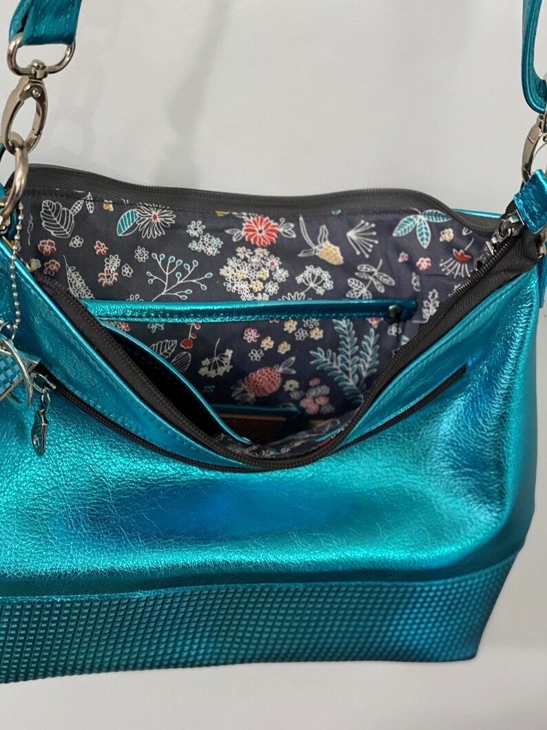 close up of turquoise Blue shiny handbag with floral patterned interior on a light gray background. slip pocket and easy access zipper closure